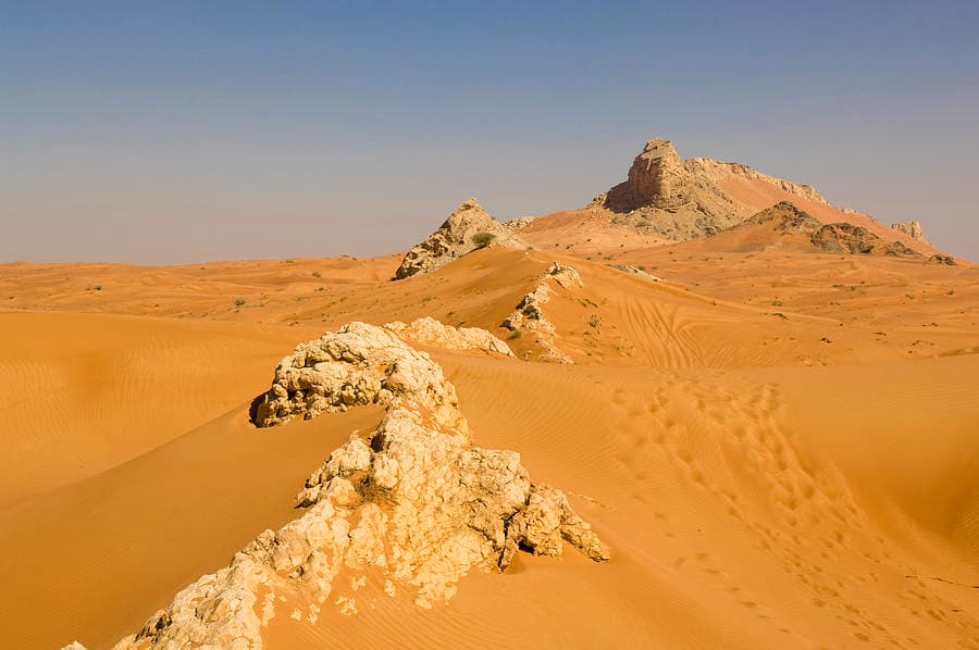 Wide desert landscape featuring golden sand dunes and rocky outcrops under a clear blue sky, characteristic of the Mleiha region.