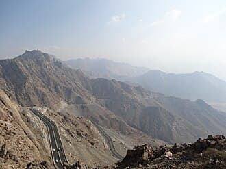 A winding paved road snakes through rugged, arid mountains under a hazy sky, capturing the dramatic landscape of the Ta'if valley.