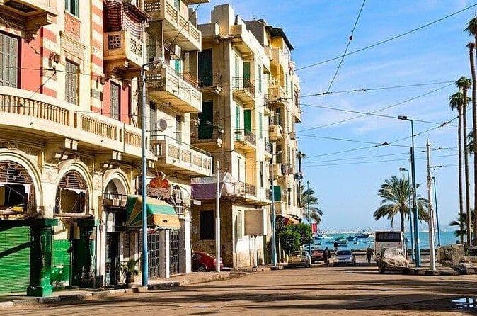 Street scene with colorful old-town buildings, balconies, palm trees, and a view of the sea in the background.