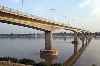 A wide-angle view of a concrete bridge spanning the Mekong River at Thakhèk, featuring sturdy pillars rising from the calm water under a clear sky.