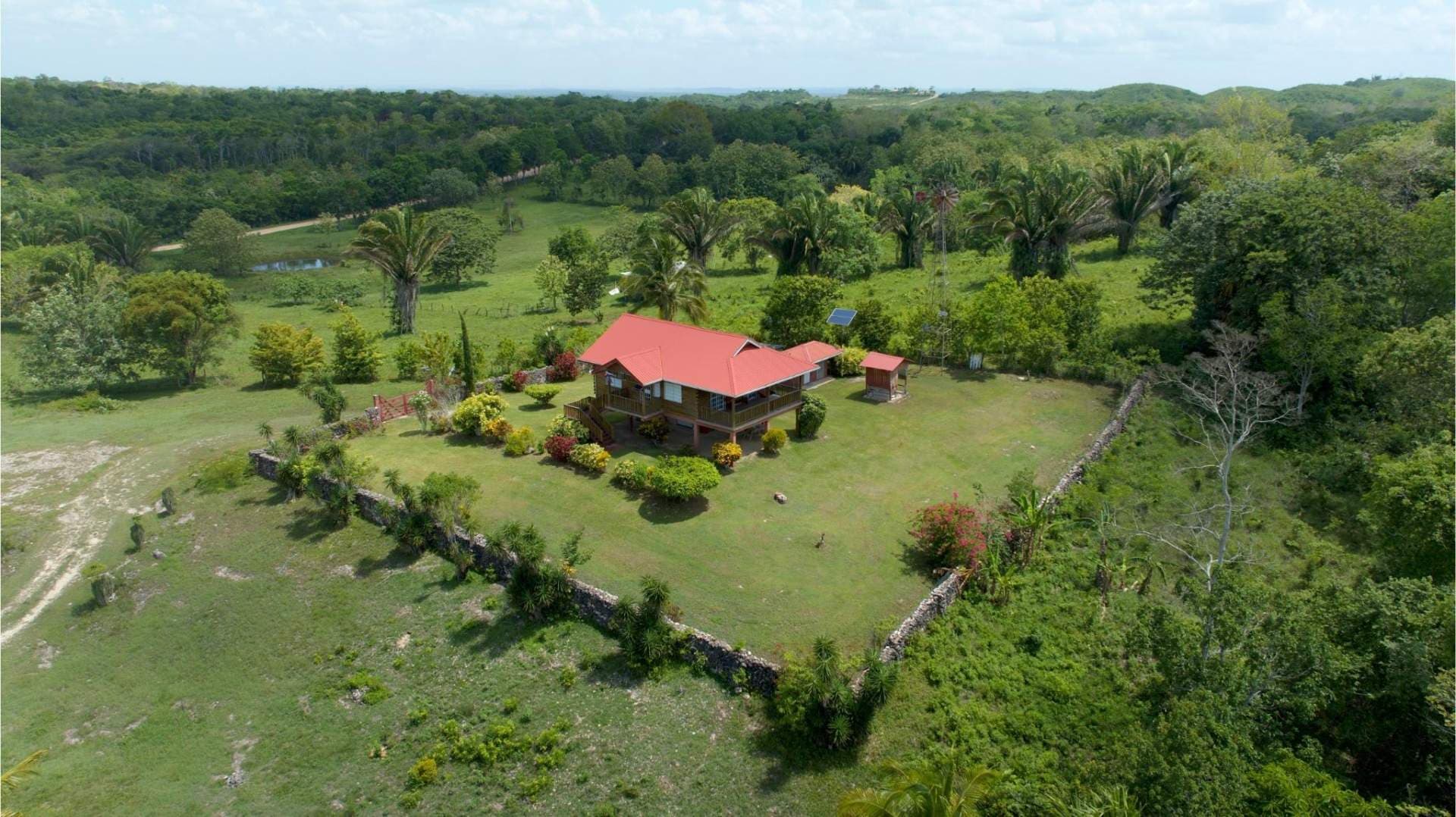 Aerial view of a red-roofed house on a green lawn surrounded by tropical trees and rolling hills.