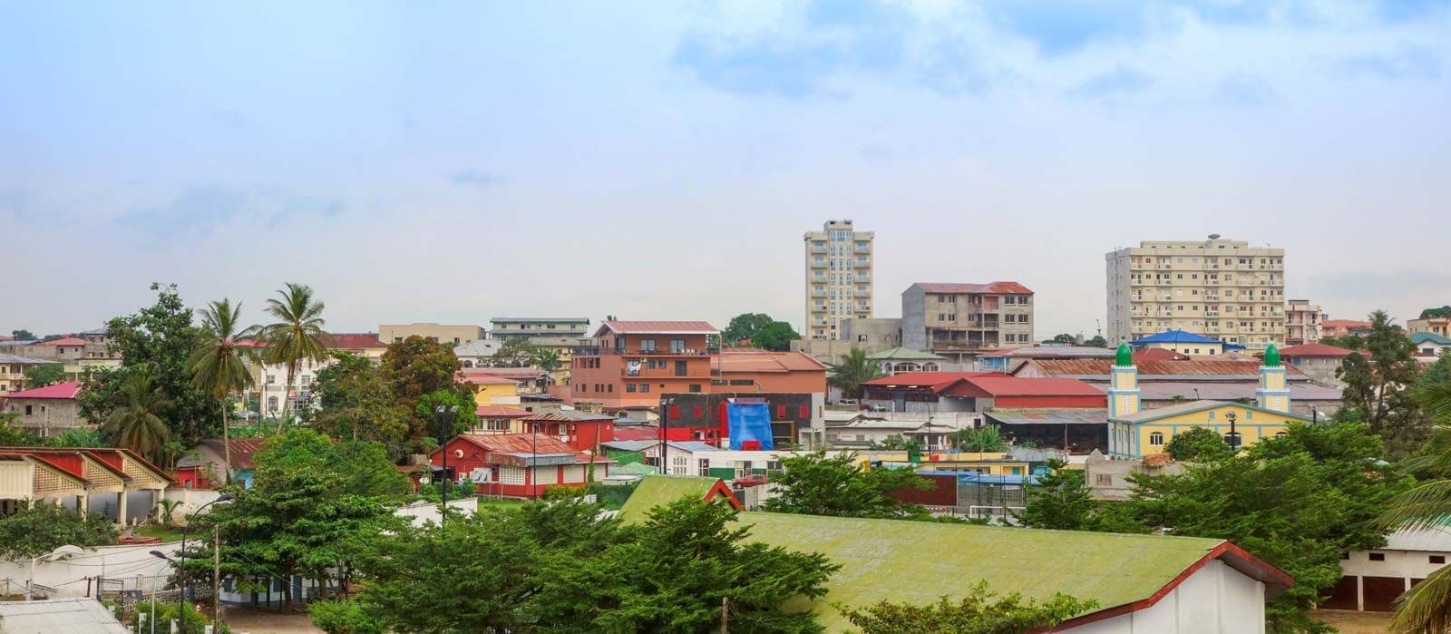 Wide-angle view of a coastal town skyline with low-rise buildings, palm trees, and a green-domed church under a partly cloudy sky.
