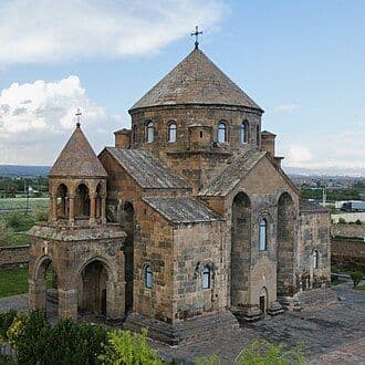 Exterior of a historic stone Orthodox church with a central dome and bell tower, set against a partly cloudy sky.