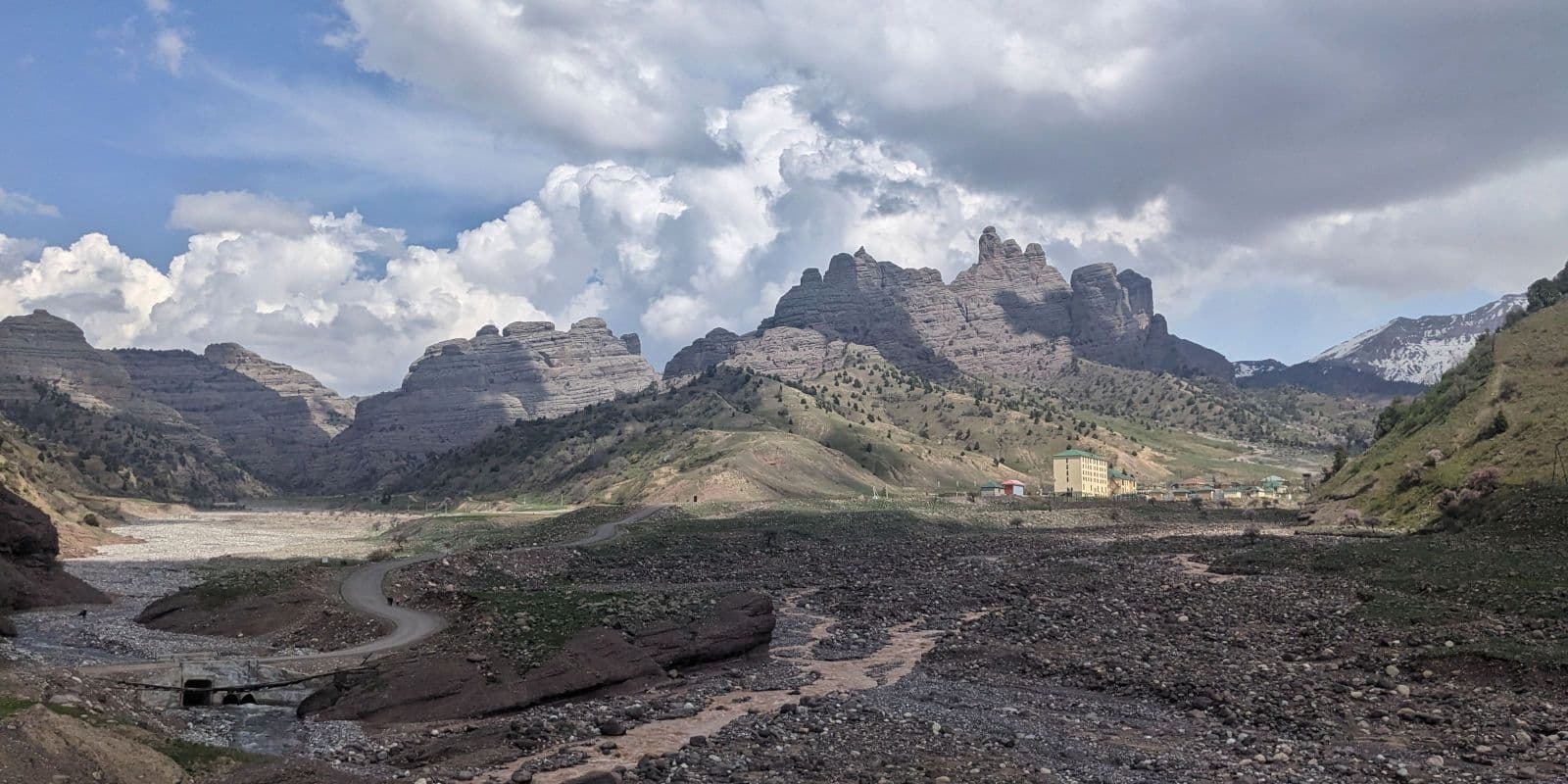 Arid landscape in Khatlon Province featuring a winding road, rocky riverbed, and dramatic stratified mountains under a cloudy sky.