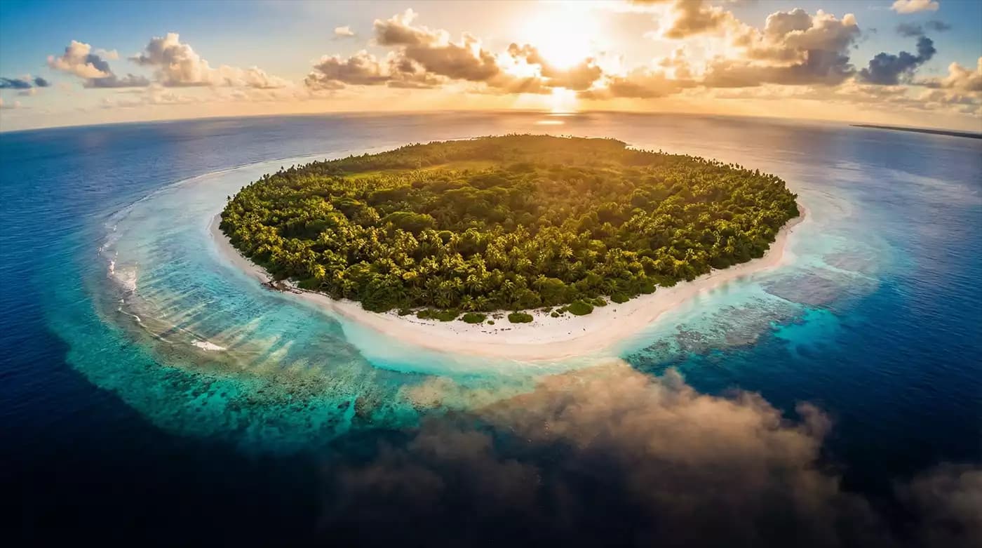 Aerial view of a small tropical island with dense green vegetation, white sand beach, and turquoise lagoon at sunset.
