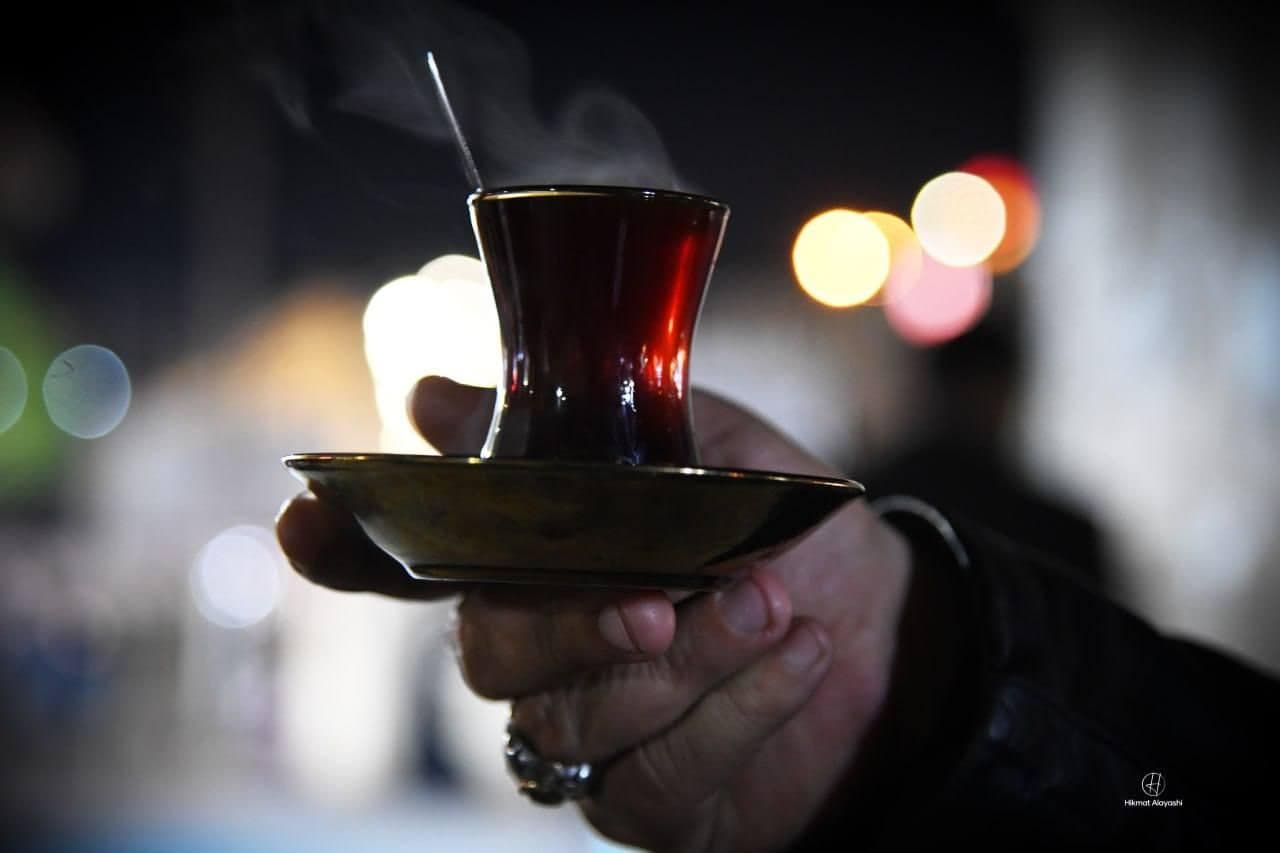 A hand holds a small glass cup of hot tea on a saucer with steam rising, a symbol of Iraqi hospitality.