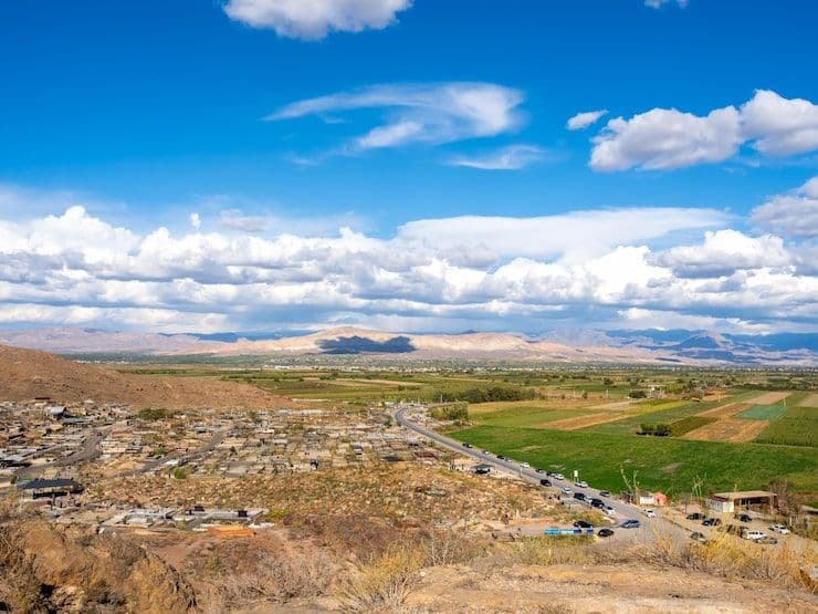 Wide landscape photograph showing a small settlement, agricultural fields, and distant mountains under a bright blue sky with scattered clouds.