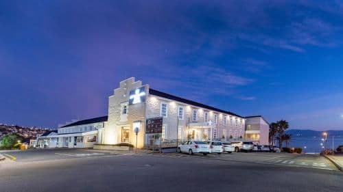 Wide-angle twilight photograph of a large, illuminated building with a cross sign, parked cars, and a coastal view.