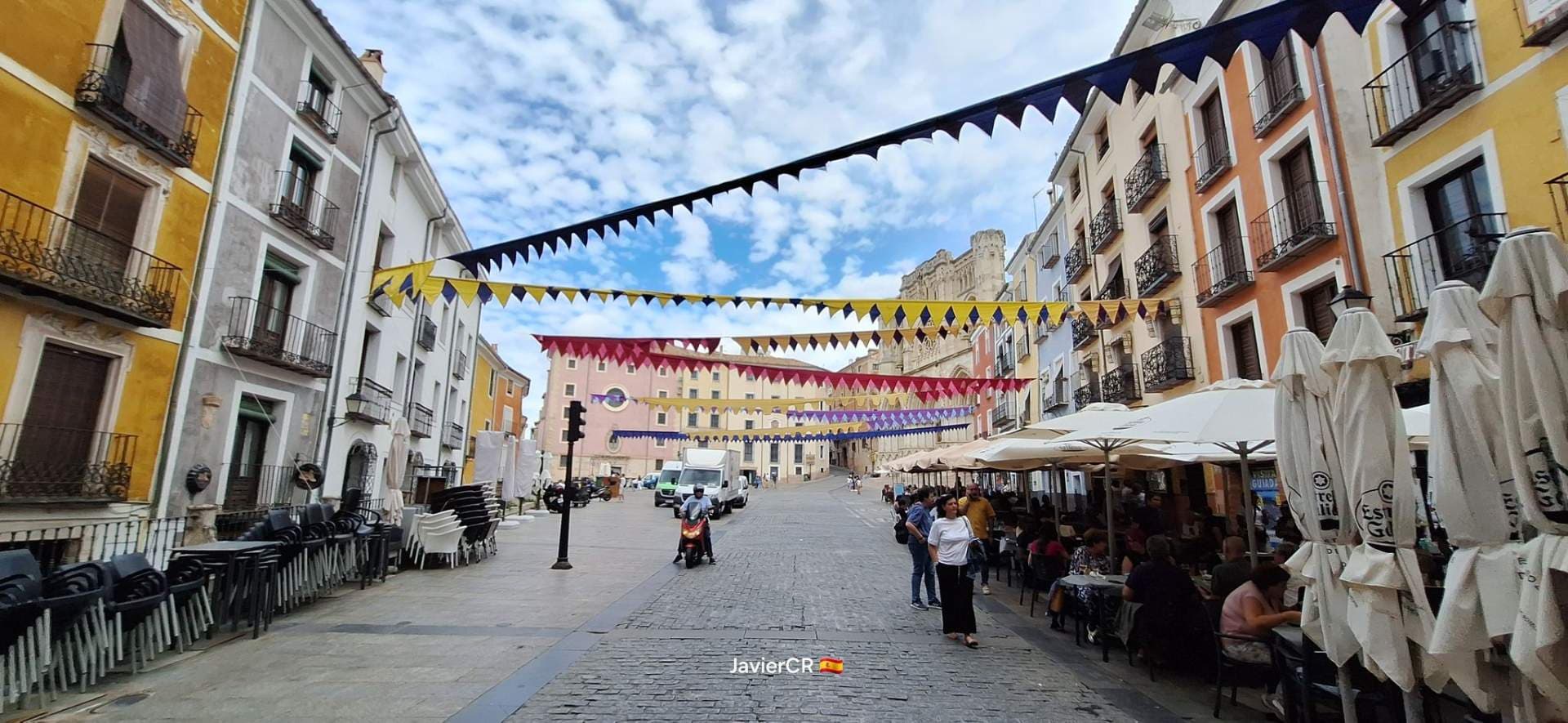 Wide street scene in a Spanish town square with colorful bunting, café umbrellas, pedestrians, and a church facade in the distance.