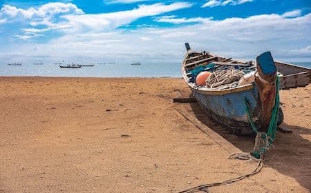 Weathered fishing boat beached on the sandy shoreline of the Anloga lagoon with distant vessels under a blue sky.