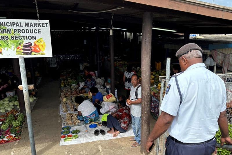 A bustling municipal market scene in Labasa featuring vendors selling fresh produce on the ground, with a security guard standing in the foreground.