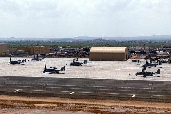Aerial view of a small regional airport runway with parked helicopters, hangars, and distant hills under a cloudy sky.
