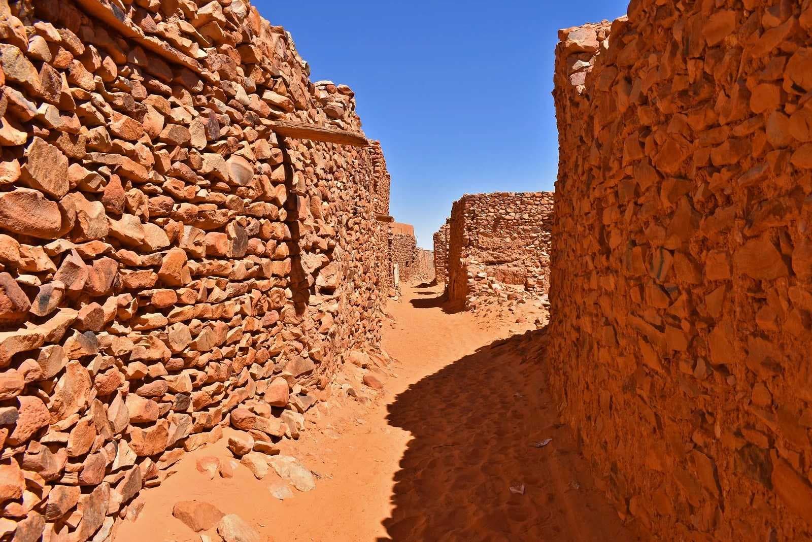 Narrow sandy alley between tall reddish earthen stone walls under a clear blue sky, typical of a Mauritanian town streetscape
