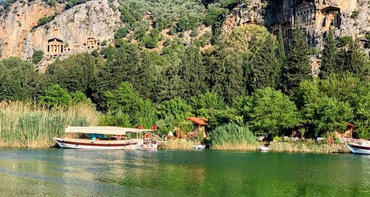 A tour boat travels along the Dalyan River, framed by tall reeds and the iconic Lycian rock tombs carved into the cliffside.