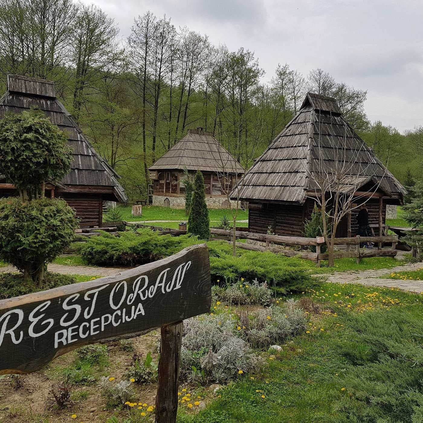 Traditional wooden log cabins with shingle roofs in a green garden, with a wooden sign reading 'RESTORANI RECEPCIJA' in Derventa.