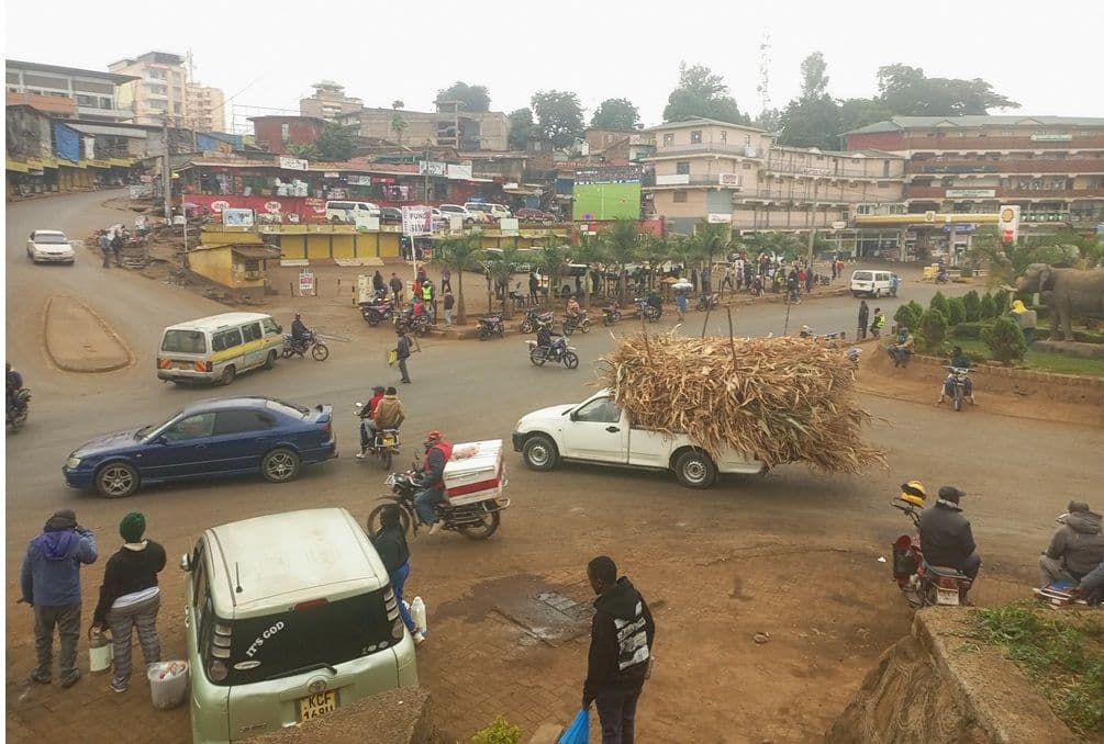 Busy town intersection in Chuka, Kenya, with cars, motorcycles, pedestrians, and a pickup loaded with dried maize stalks.