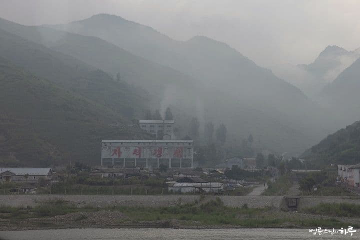 Misty mountain valley with a factory building bearing red Korean text, small village structures, and a river in the foreground.