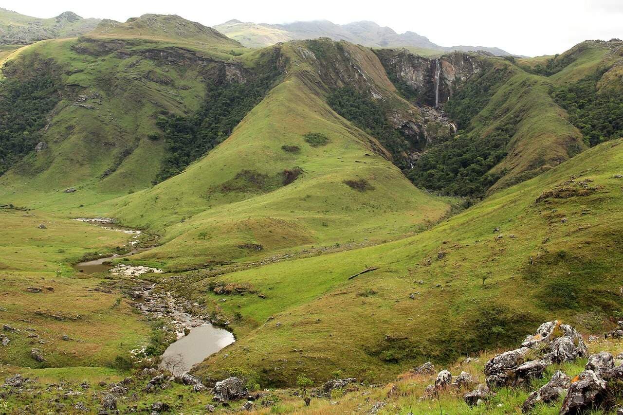 Green rolling hills with a small waterfall and stream under an overcast sky near the Chimanimani mountains.