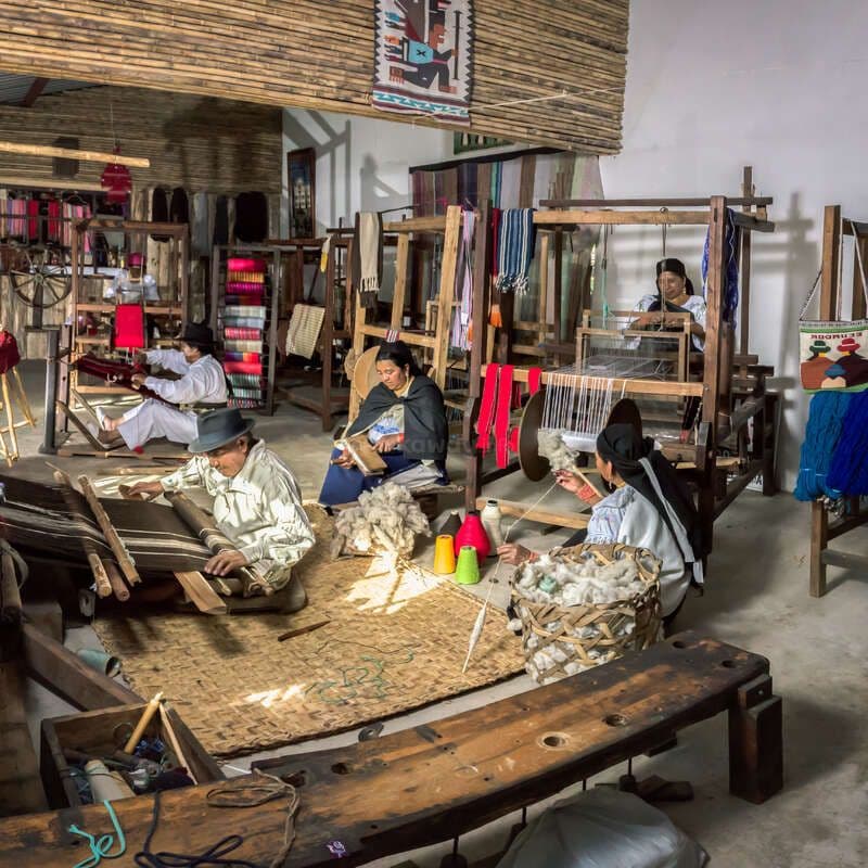 Artisans weaving textiles on traditional looms inside a workshop, surrounded by colorful yarns and finished fabrics in the Atuntaqui textile district.