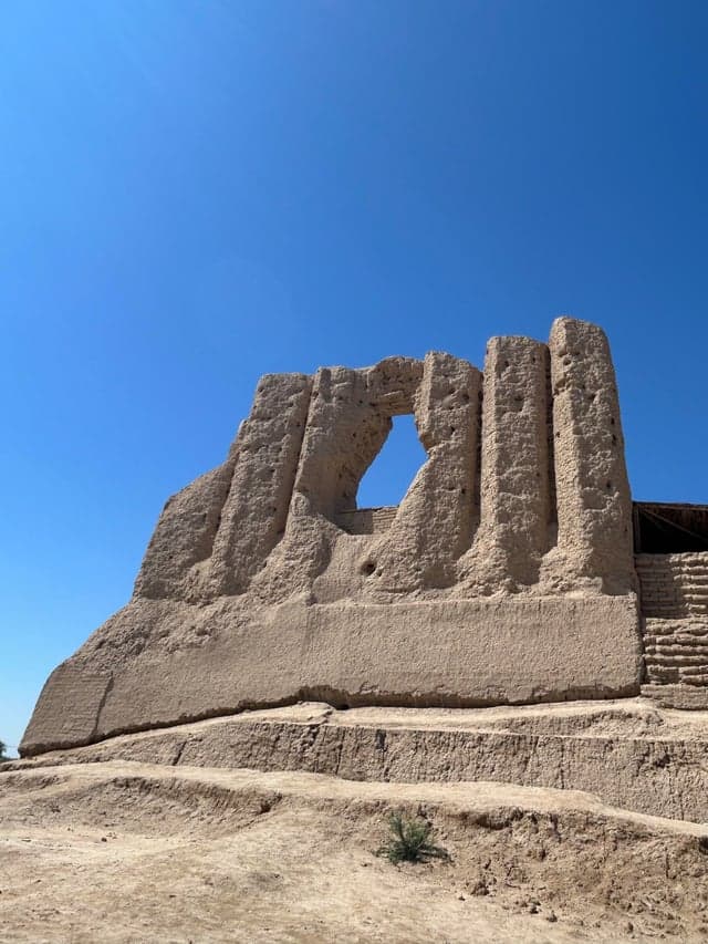 Sunlit ruins of Gokdepe Fortress featuring weathered mud-brick walls and a distinct arched opening against a clear blue sky.
