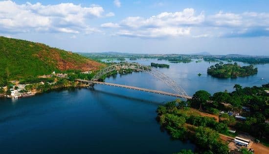 Aerial view of a large arch bridge spanning a wide lagoon near Anloga, with green hills and scattered buildings on the banks.