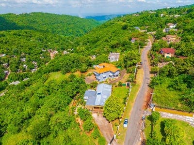 Aerial view of a winding rural road cutting through lush green hills in Saint Andrew, Jamaica, with scattered houses and a blue car visible.