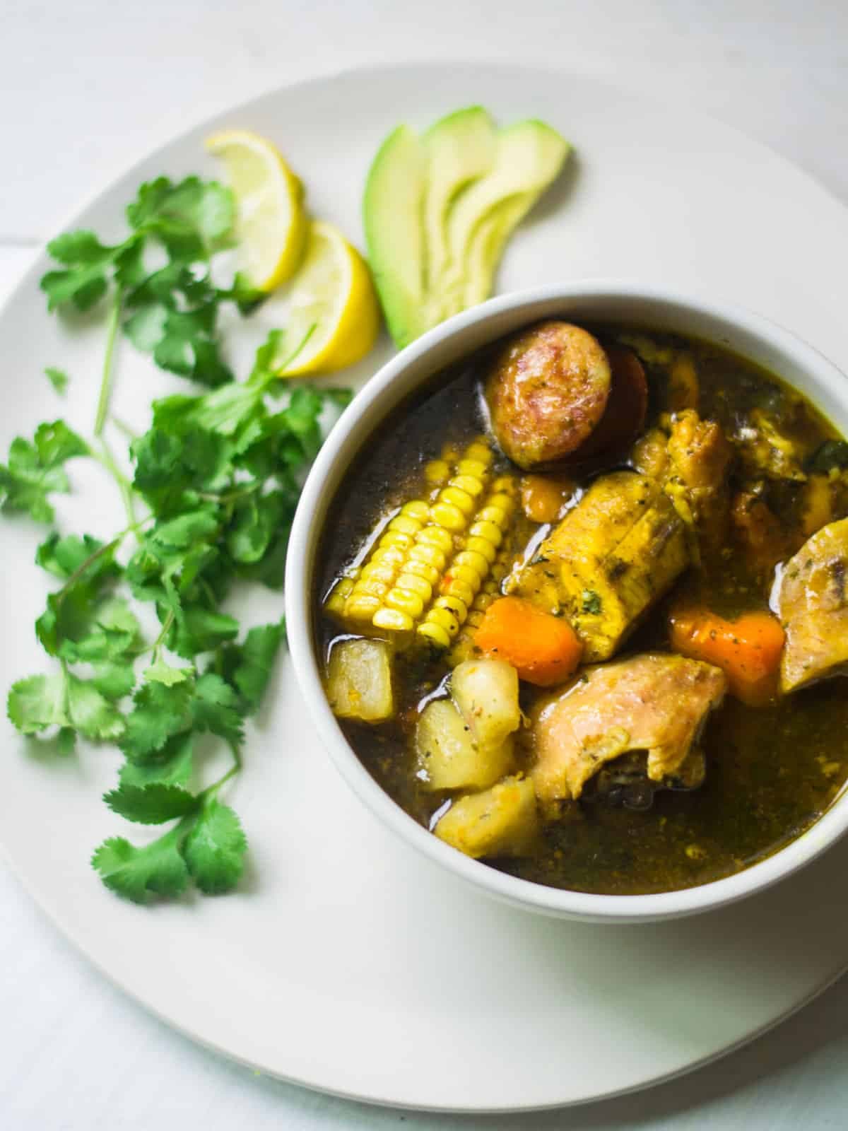 Close-up of a bowl of traditional Dominican sancocho stew with corn, potatoes, and meat, served with cilantro, lemon, and avocado.