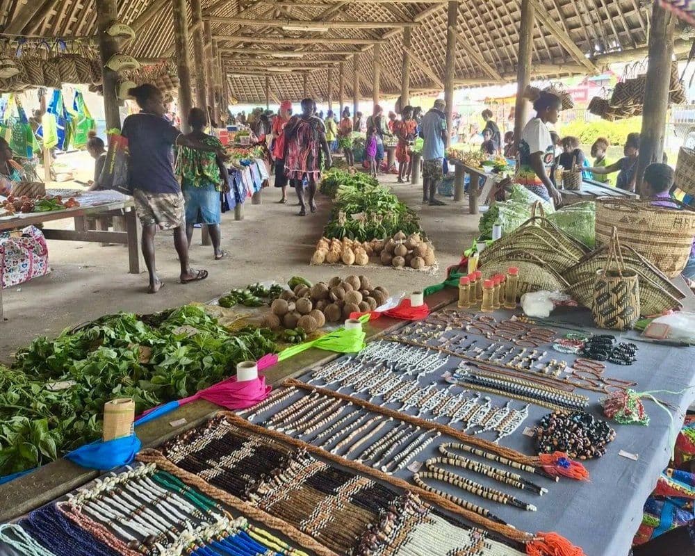 Open-air market with stalls displaying fresh produce, woven baskets, and beadwork under a thatched roof