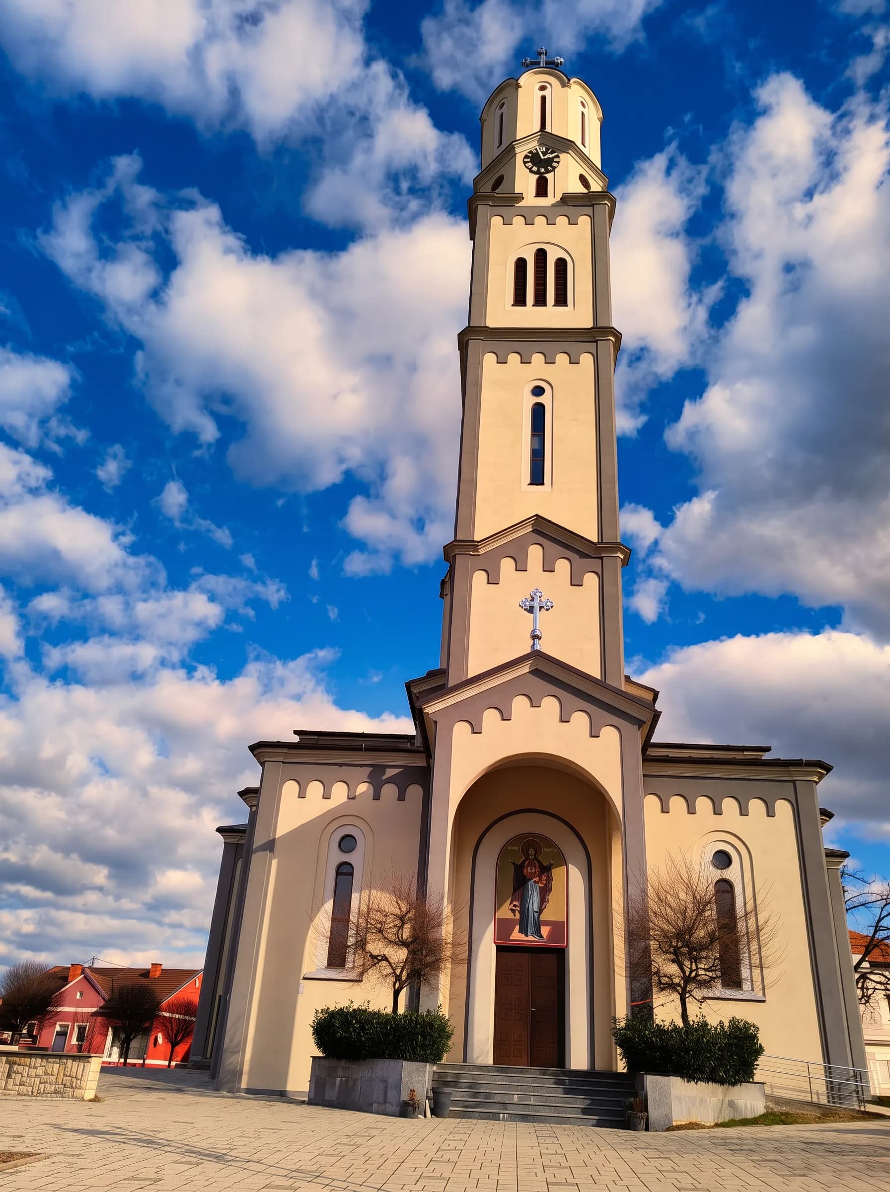 A tall beige church tower with a clock and cross dominates the frame, set against a blue sky with clouds in Derventa.