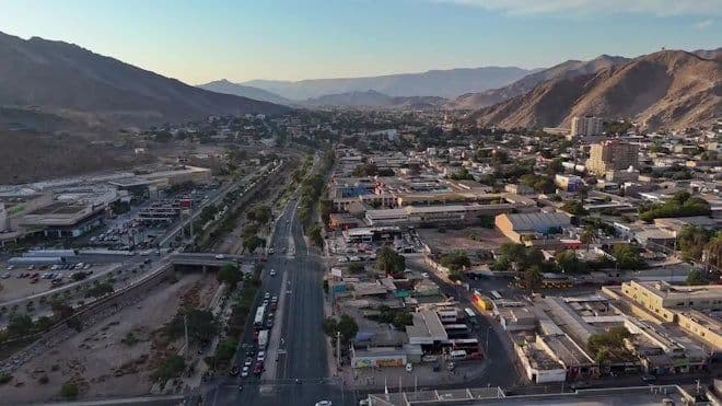Aerial view of a major avenue cutting through a desert city flanked by mountains, with buildings and traffic visible