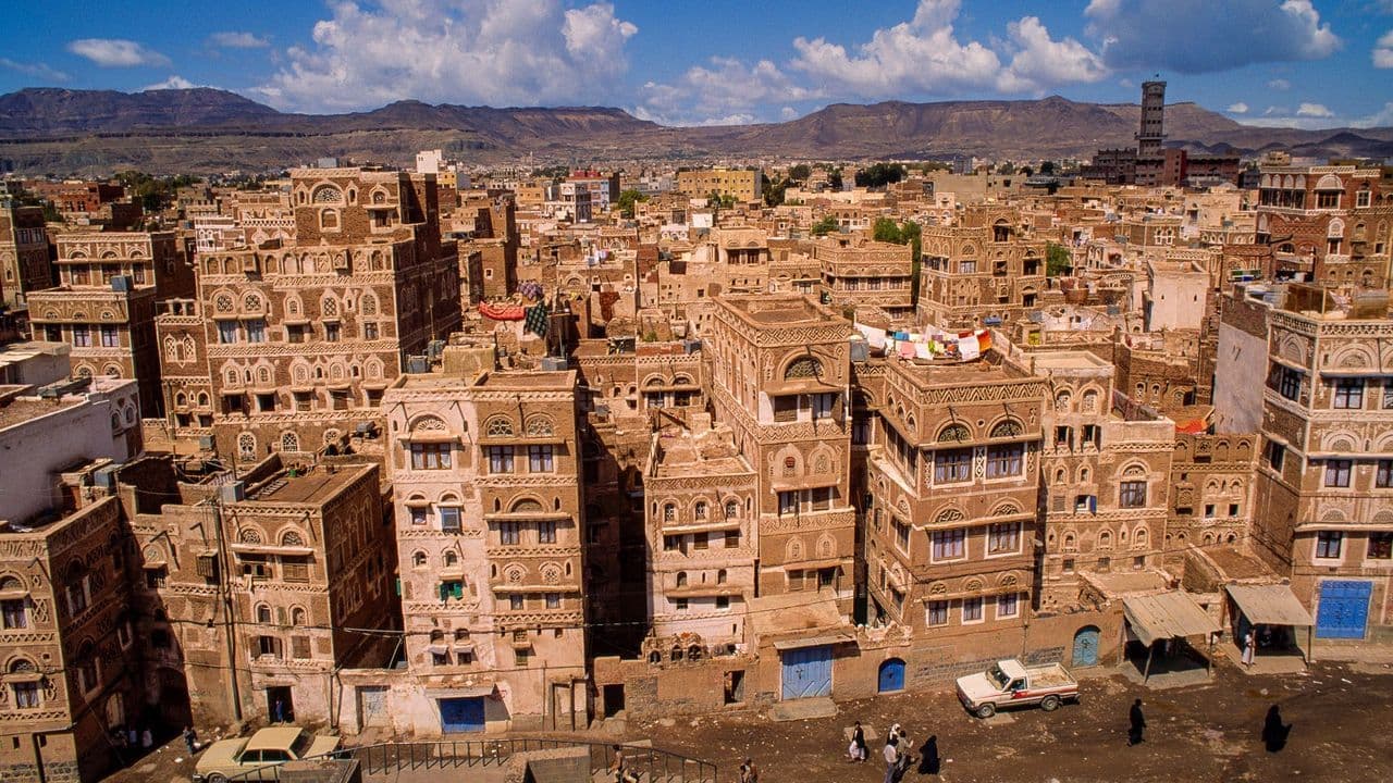 Aerial view of traditional Yemeni mudbrick architecture in a rural highland village near Wadi Al Ayn, with mountains in the background.