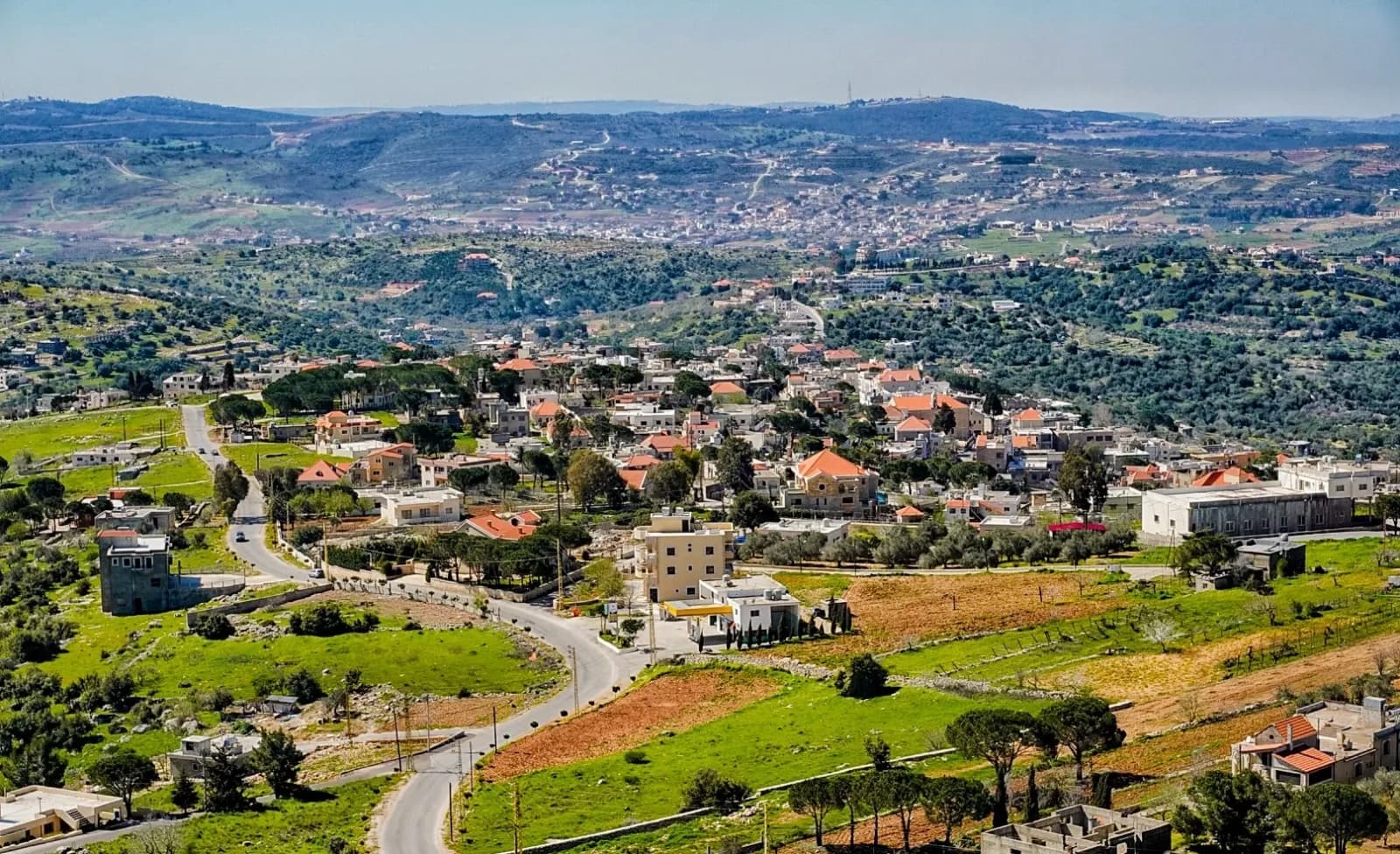 Aerial panoramic photograph of a hillside village with red-roofed buildings, winding roads, green fields, and distant hills under a clear sky.