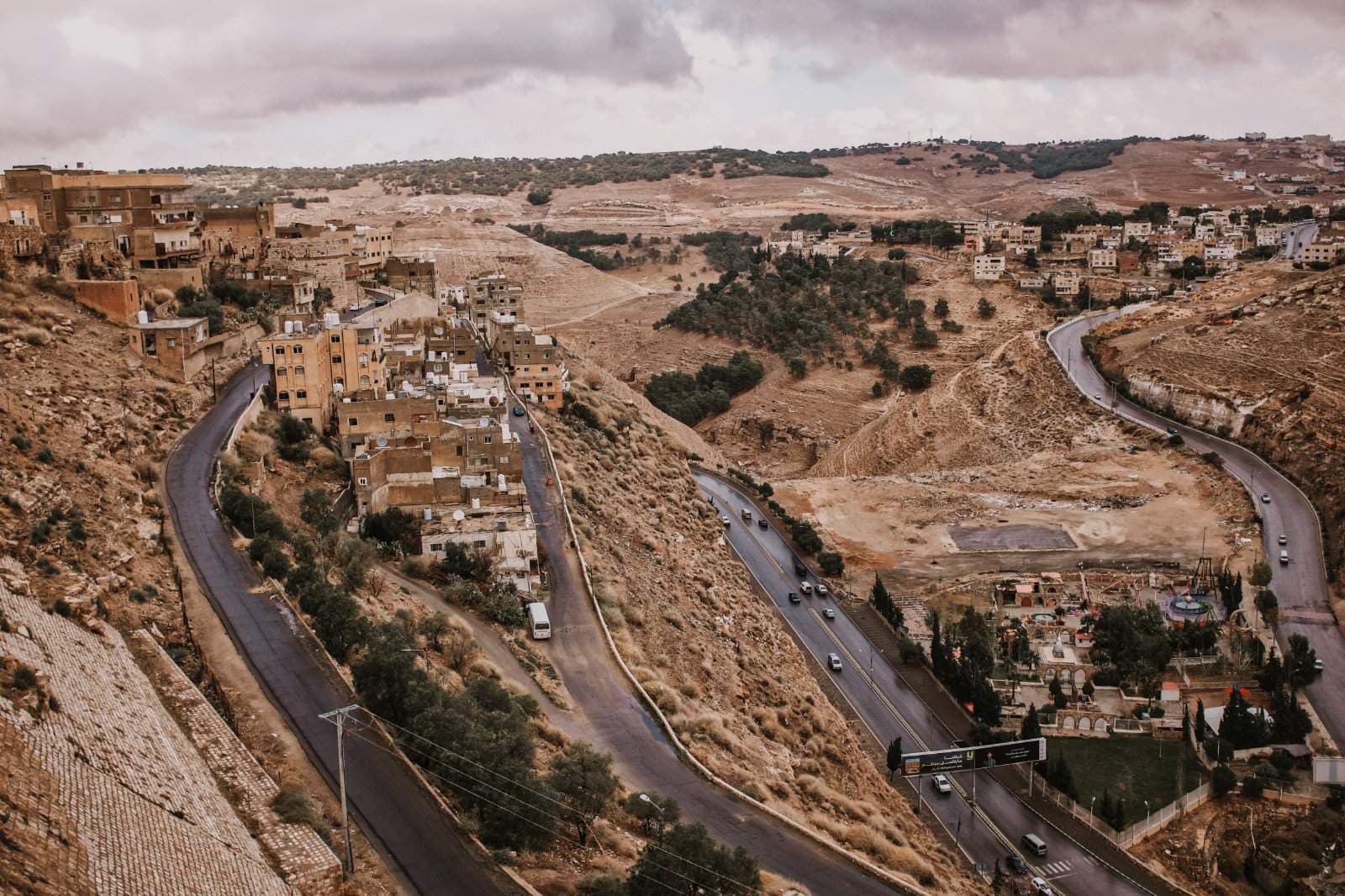 Aerial view of winding roads cutting through arid, hilly terrain with clustered stone buildings and sparse vegetation under a cloudy sky.