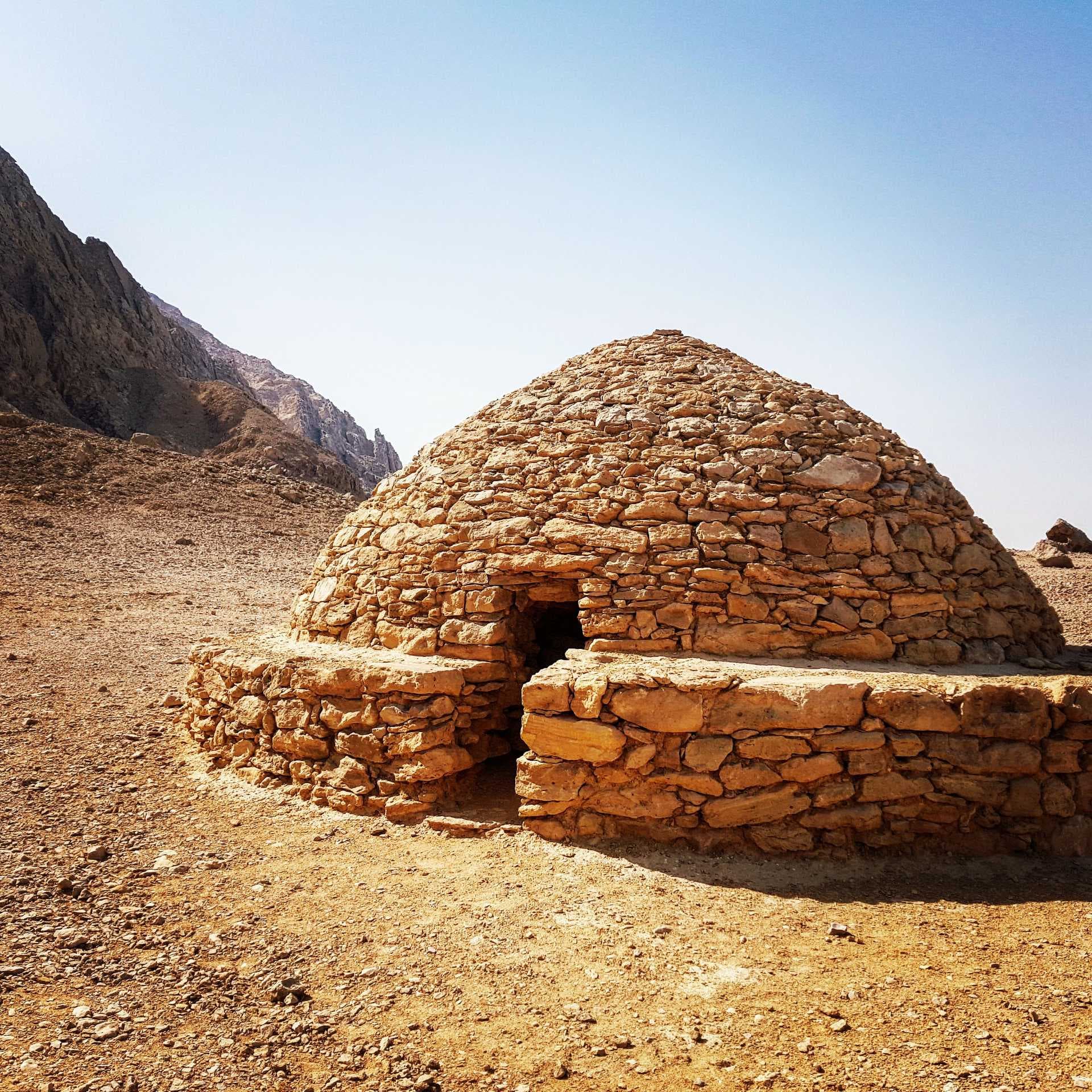 Ancient stone beehive tomb structure situated in the arid Mleiha desert landscape with rocky mountains in the background under a clear blue sky.