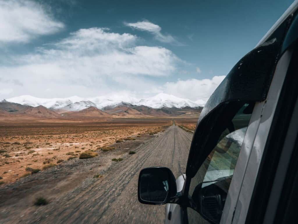 View from a vehicle on a dusty road in a desert landscape with snow-capped mountains in the distance.