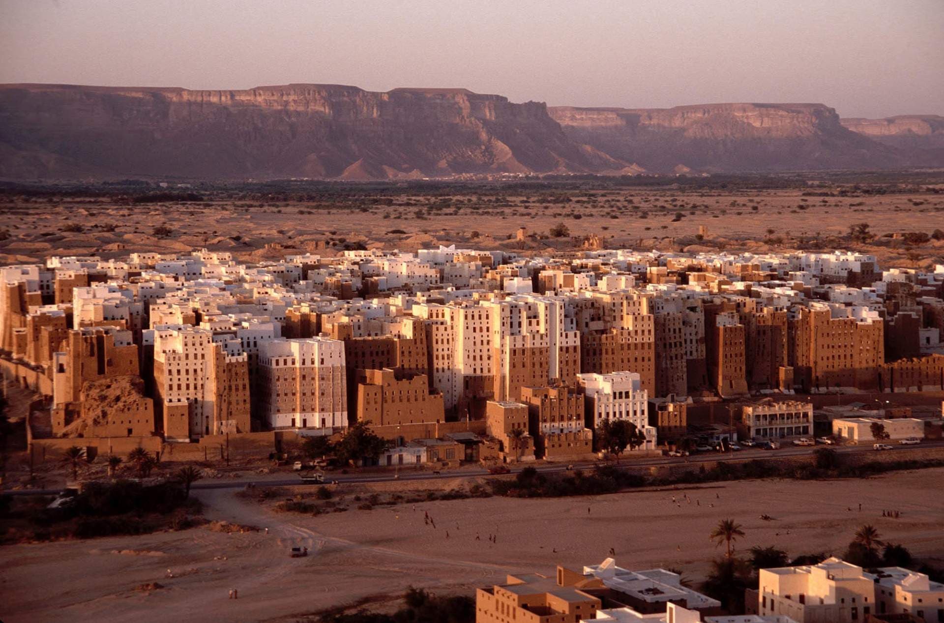 Aerial view of As Sawm village featuring traditional mud-brick tower houses against a backdrop of desert plains and distant mountains.