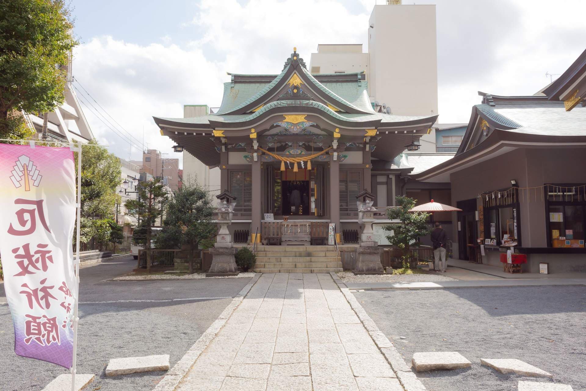 Traditional Japanese shrine building with green roof and stone lanterns set on a paved street with surrounding urban structures