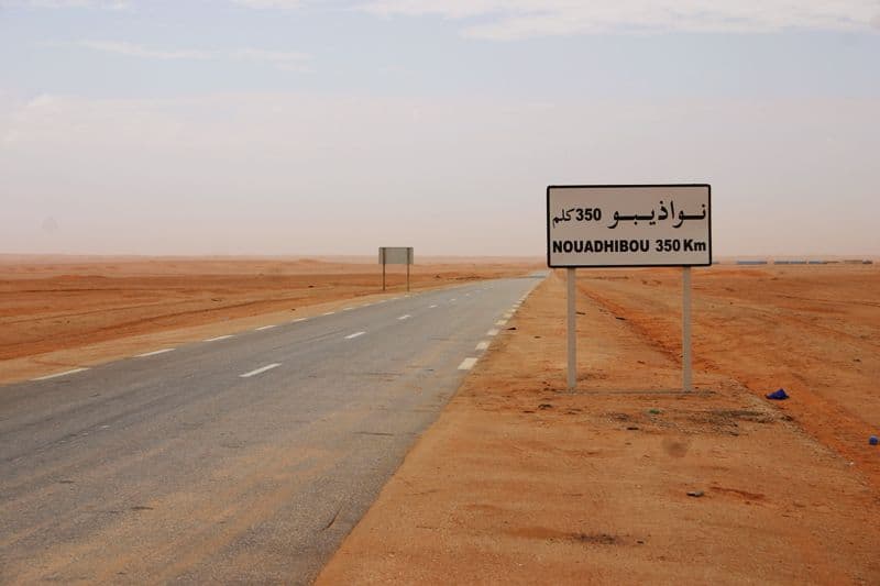 Paved highway stretches through arid desert landscape with a road sign indicating Nouadhibou 350 km ahead in Mauritania