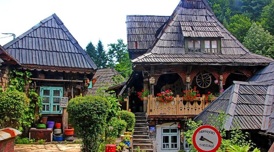 Traditional wooden houses with steep shingled roofs and flower-adorned balconies at the Ethno Village in Banovići, surrounded by lush greenery.
