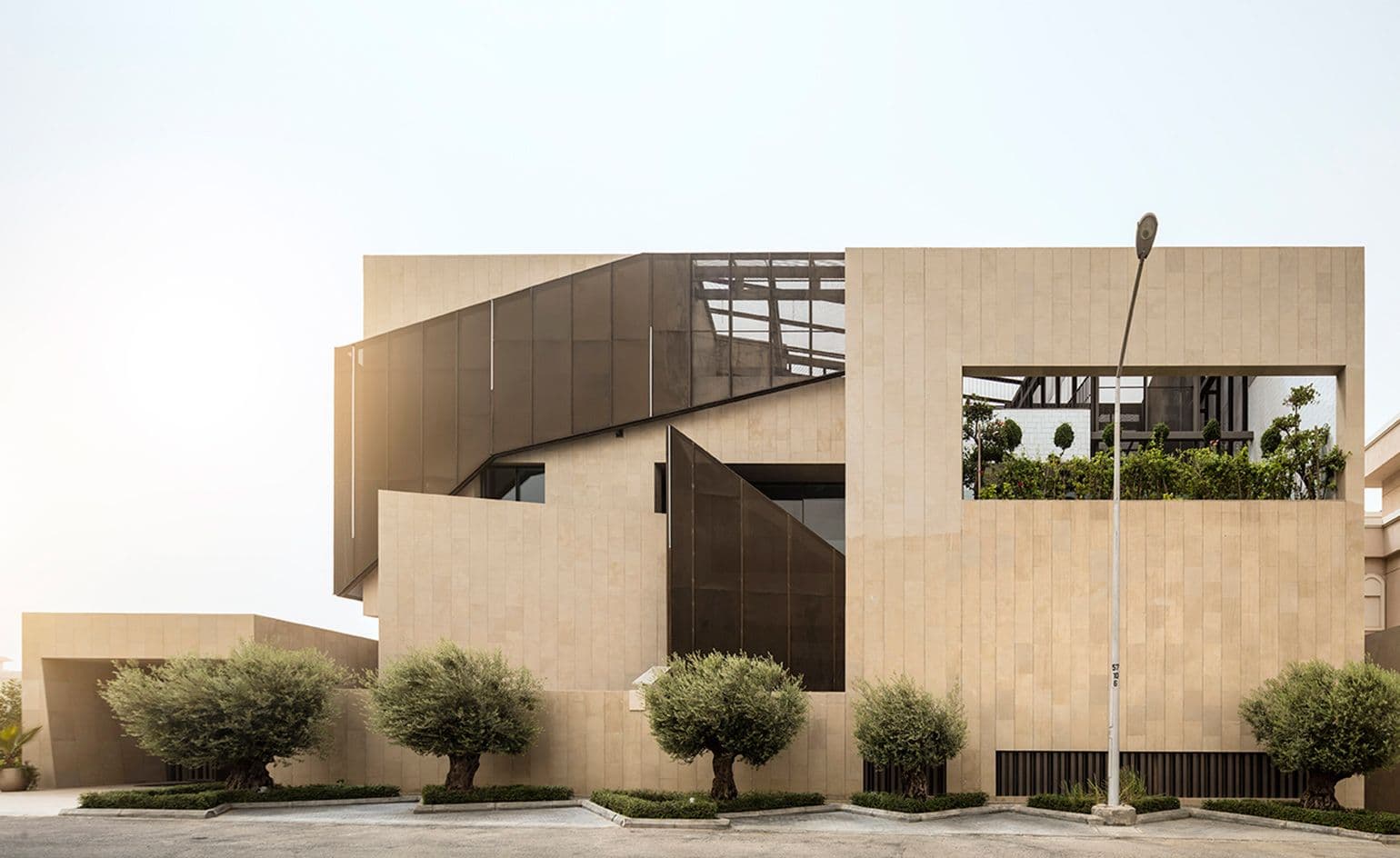 Wide exterior of a modern residential building in a Kuwait suburb with beige stone walls, dark glass panels, and landscaped olive trees