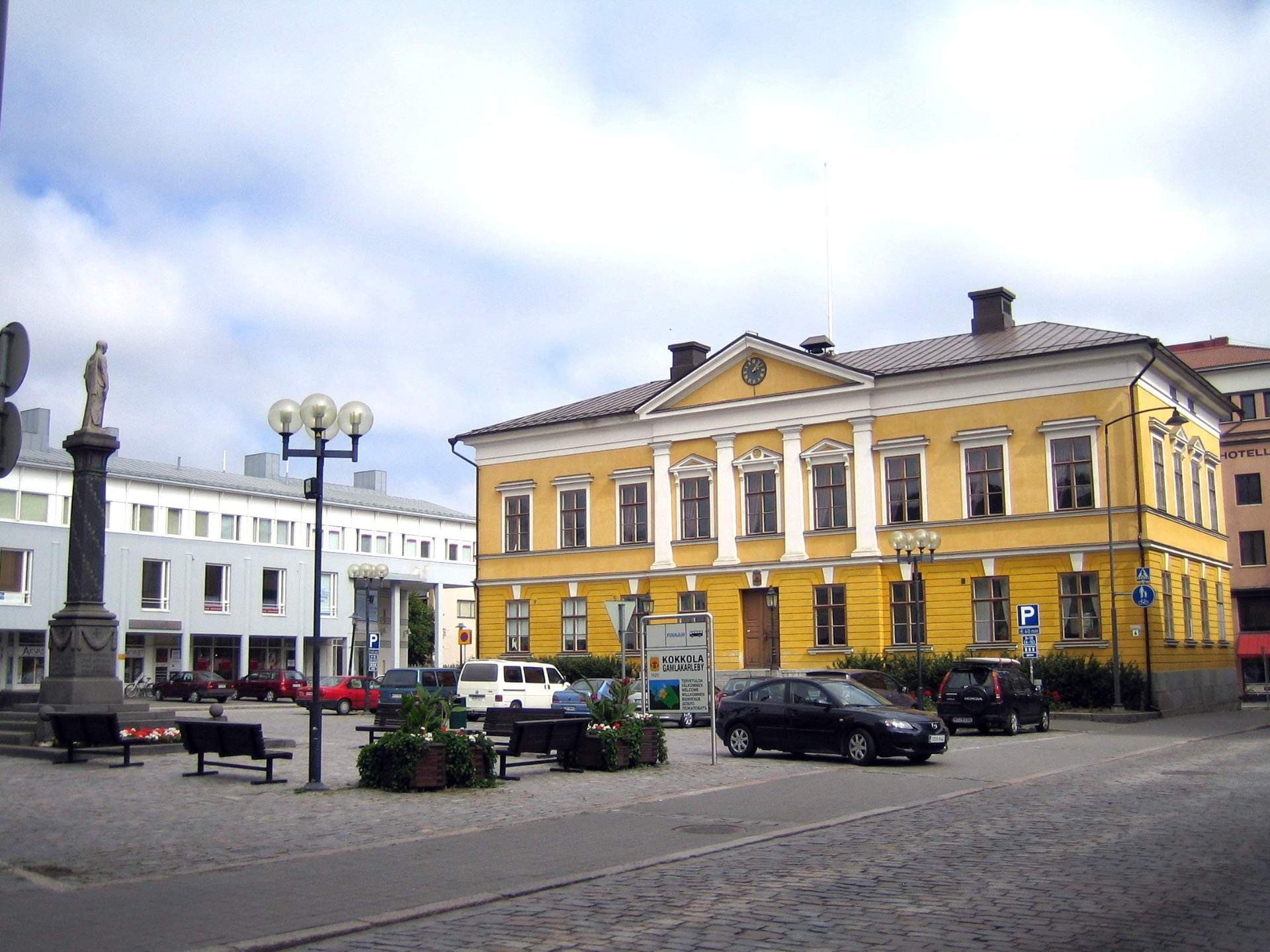 A yellow classical building stands in a cobblestone town square with a statue, benches, and parked cars under a cloudy sky.