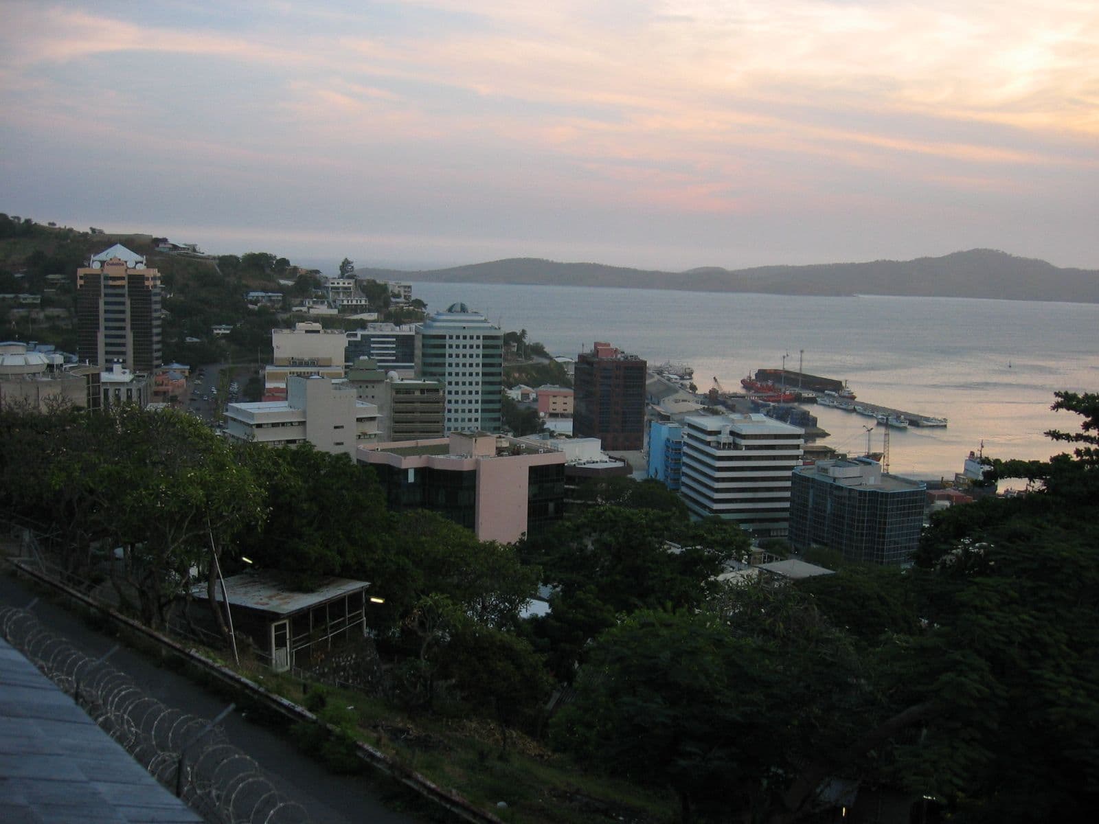 Aerial view of a coastal city at dusk, showing modern buildings, a harbor with ships, and distant hills.