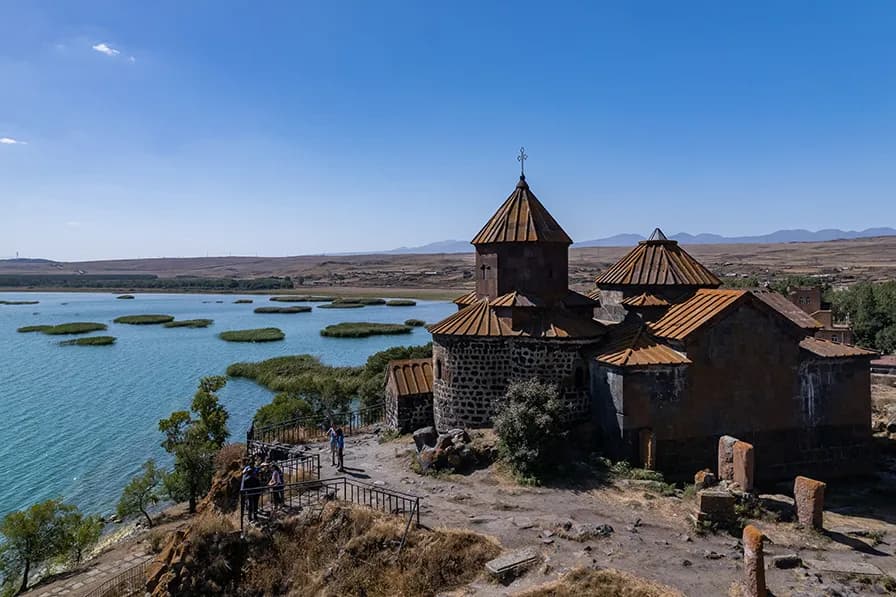 Stone church with rusted roofs beside a lake, overlooking a wide plain and distant mountains under a clear blue sky.