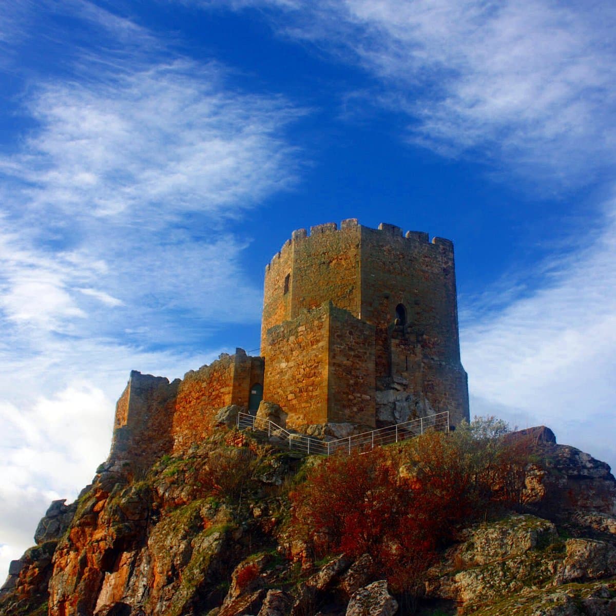 Sunlit stone tower of the Castle of Vimioso perched on a rocky hilltop against a blue sky with wispy clouds.