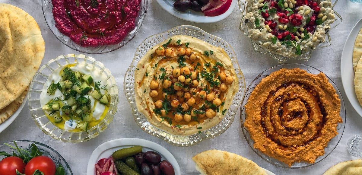 Overhead view of a Lebanese mezze spread featuring hummus, baba ganoush, tabbouleh, pickles, and flatbread on a white tablecloth.