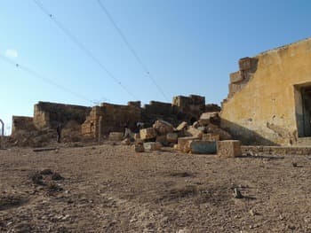 Sunlit ruins of stone and mud-brick buildings in a dry, dusty landscape under a clear blue sky with overhead power lines.