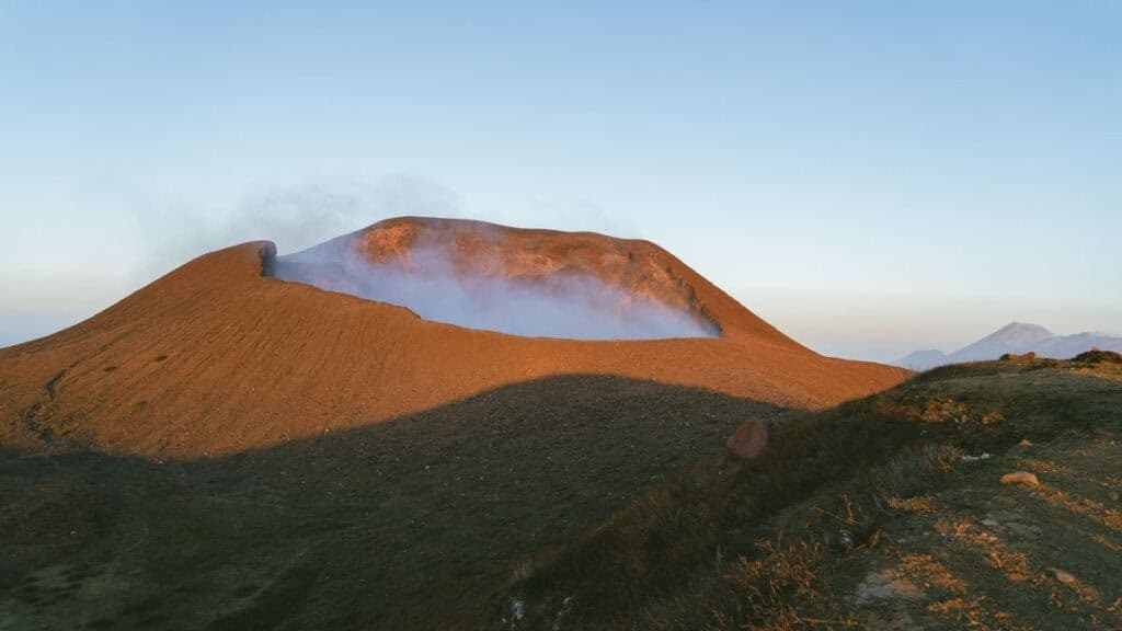 Aerial view of a smoking volcanic crater with reddish-brown slopes under a clear sky.
