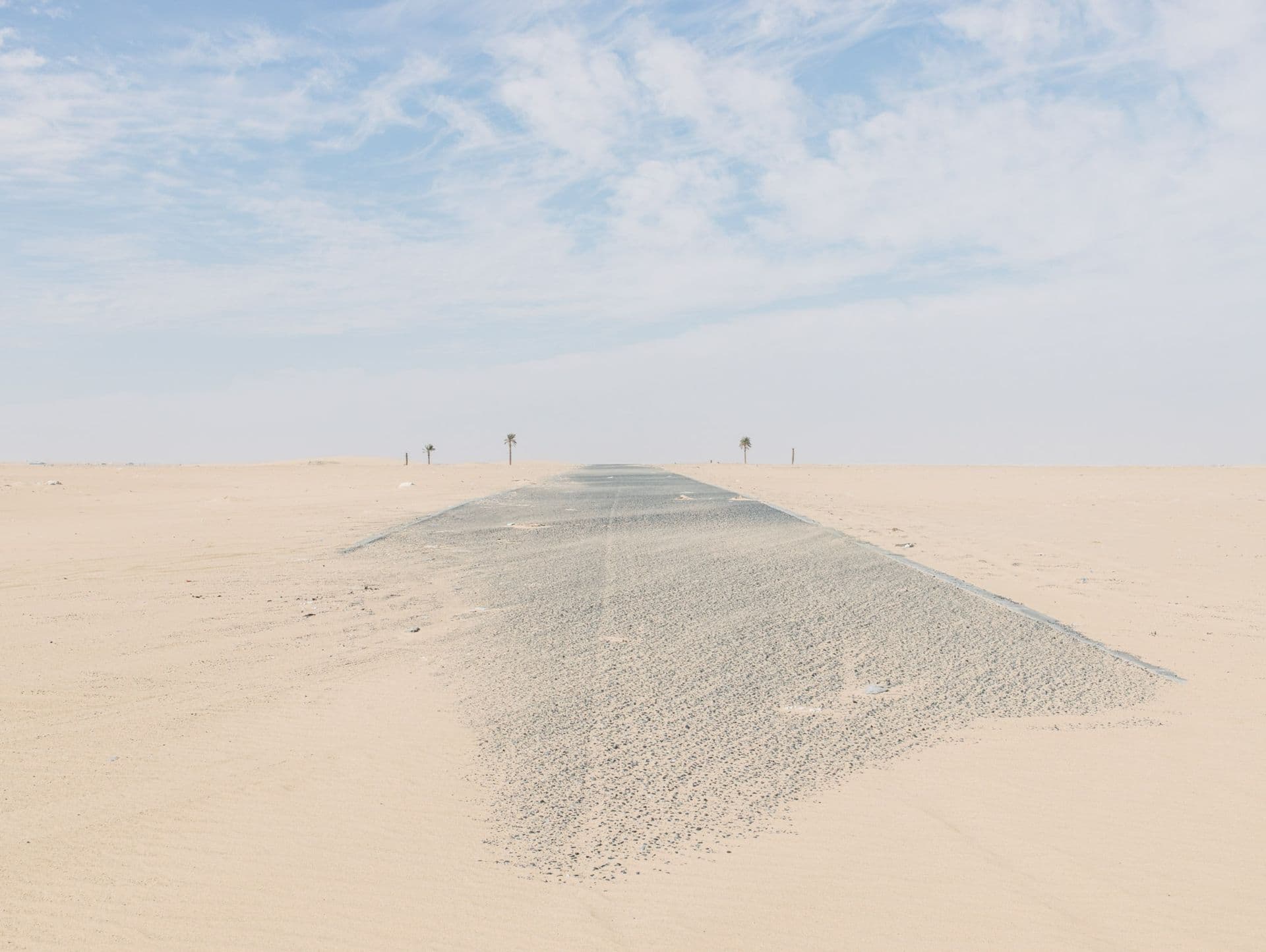 A paved road stretches through a vast, sandy desert landscape under a blue sky with wispy clouds, flanked by distant palm trees.