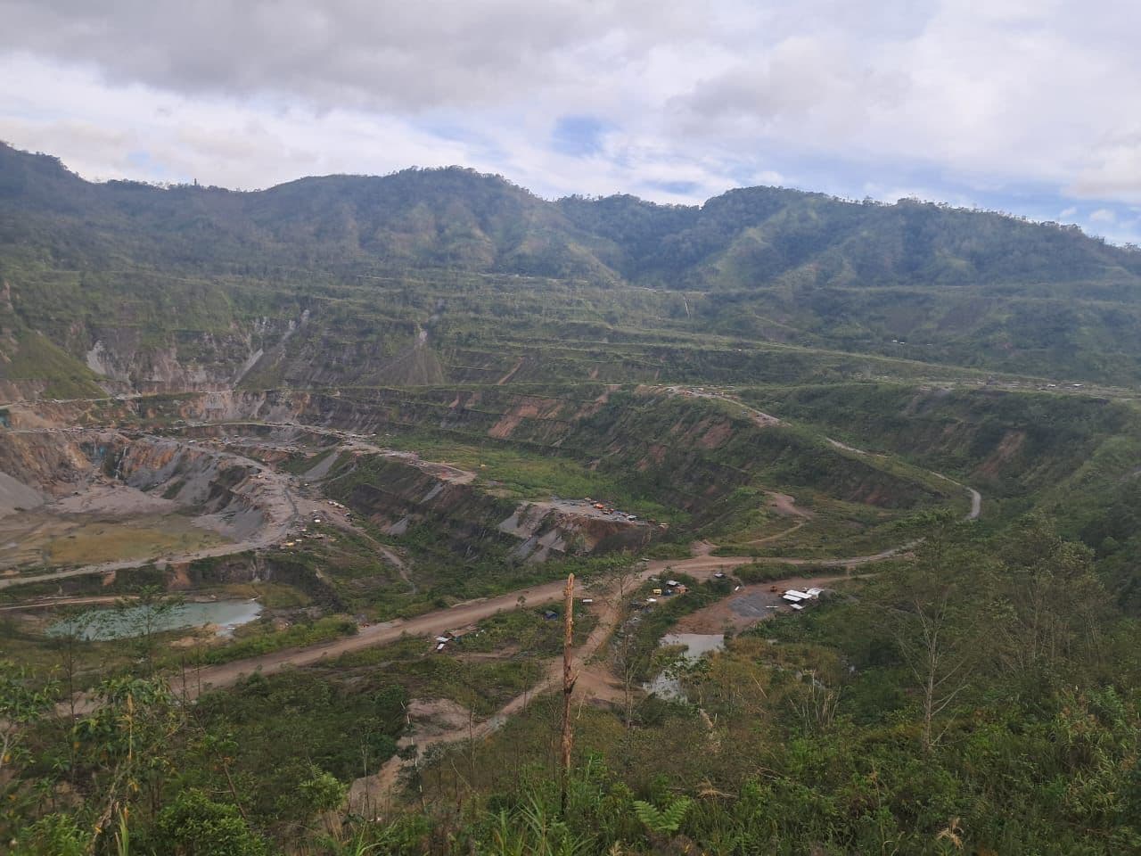 Wide aerial view of the Panguna open-pit mine with terraced slopes, dirt roads, and small structures in a mountainous landscape