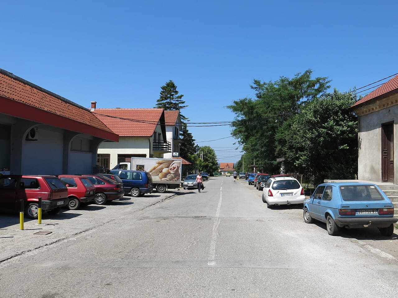 A sunny residential street in Stepojevac features parked cars, a bakery truck, and traditional buildings under a clear blue sky.
